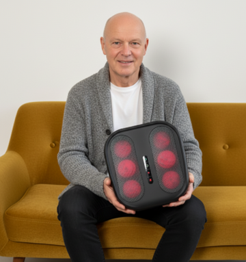 Man holding a black foot massager with red lights on a yellow couch.